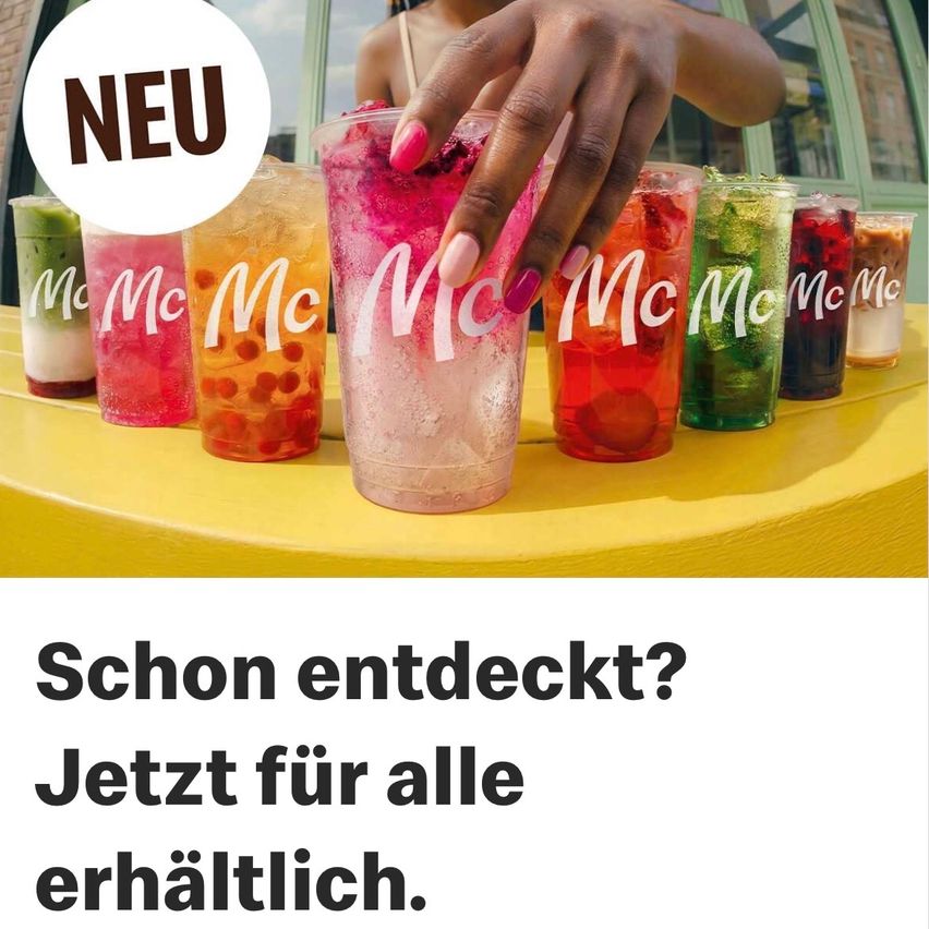 Eine Hand hält ein leuchtend pinkes Getränk von McDonald's, mit mehreren anderen bunten Getränken, die auf einem gelben Tisch aufgereiht sind. Das Wort 'NEU' ist prominent angezeigt.
