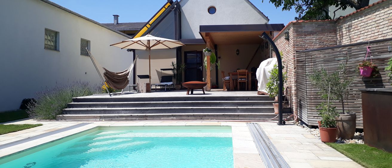 Ein modernes Haus mit Swimmingpool, Terrasse und Deck mit überdachtem Bereich, Tisch und Stühlen. Ein Schirm und eine Liege sind ebenfalls vorhanden.