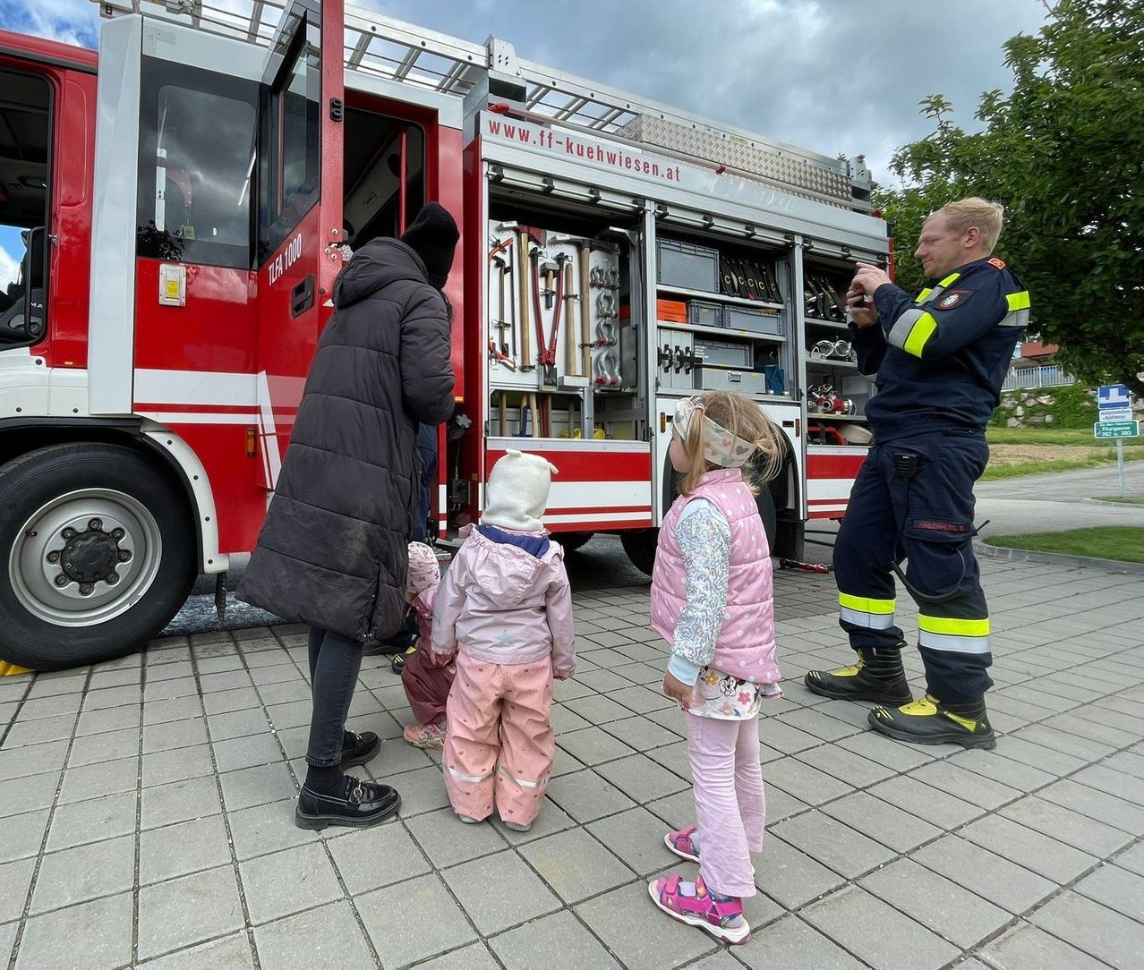 Ein Feuerwehrmann zeigt einer Gruppe von Kindern, darunter zwei Mädchen und ein Baby, das Innere eines Feuerwehrwagens.