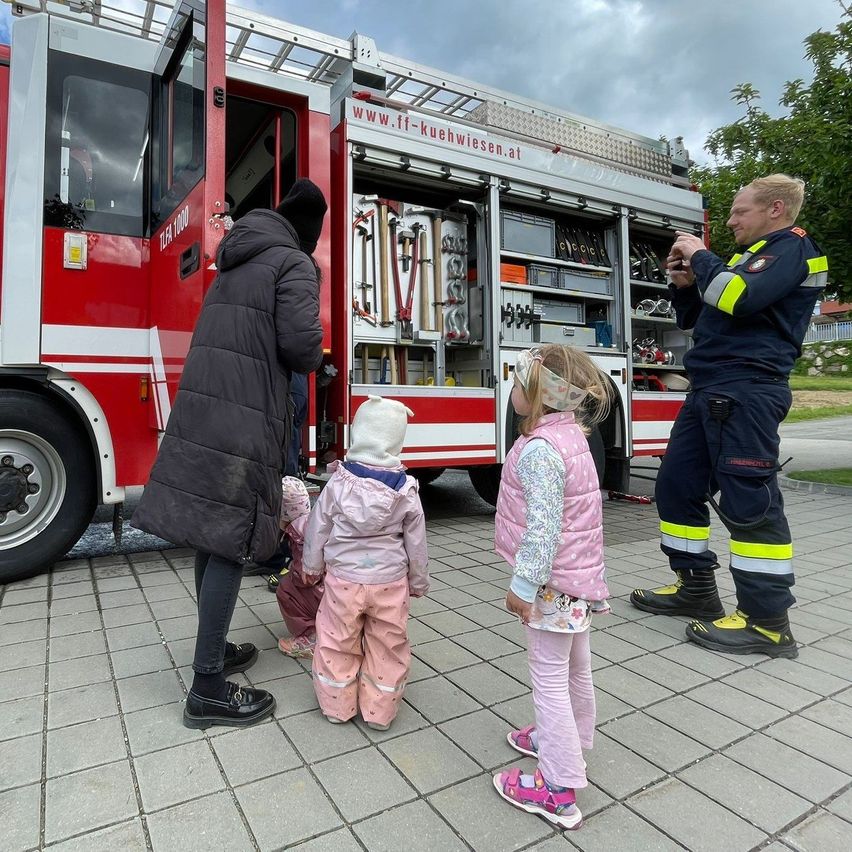 Ein Feuerwehrmann zeigt einer Gruppe von Kindern, darunter zwei Mädchen und ein Baby, das Innere eines Feuerwehrwagens.