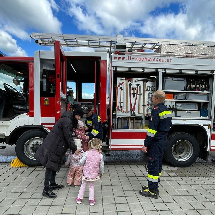Eine Frau und drei Kinder interagieren mit Feuerwehrleuten vor einem Feuerwehrwagen. Die Feuerwehrleute stehen neben den offenen Wagentüren.
