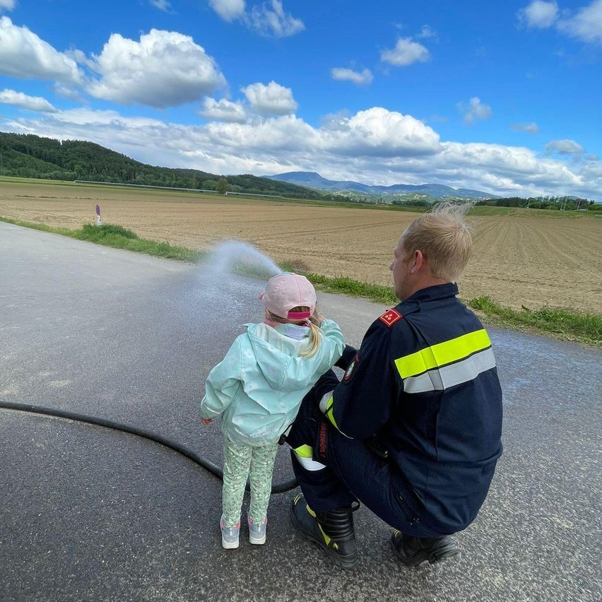 Ein Feuerwehrmann kniet neben einem kleinen Mädchen, das einen Schlauch hält und Wasser auf die Straße sprüht. Dahinter liegt ein weites Feld mit Bergen in der Ferne.