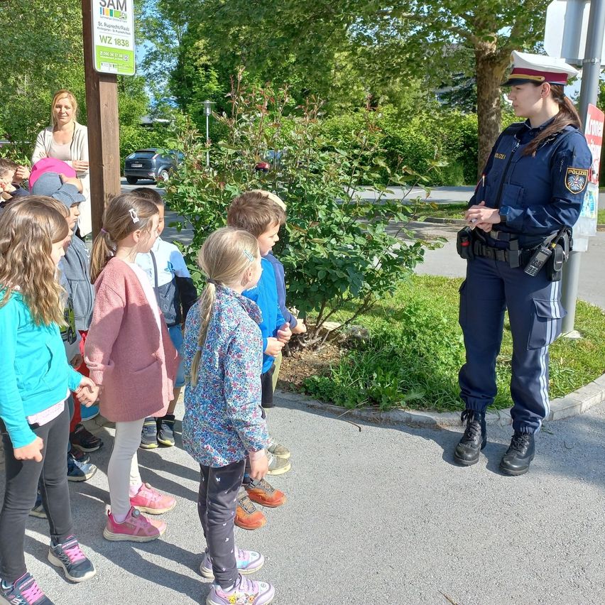 Eine Polizistin erklärt etwas einer Gruppe von Kindern auf dem Bürgersteig. In der Nähe befindet sich ein Schild mit dem Text SAM und einer Frau im Hintergrund.