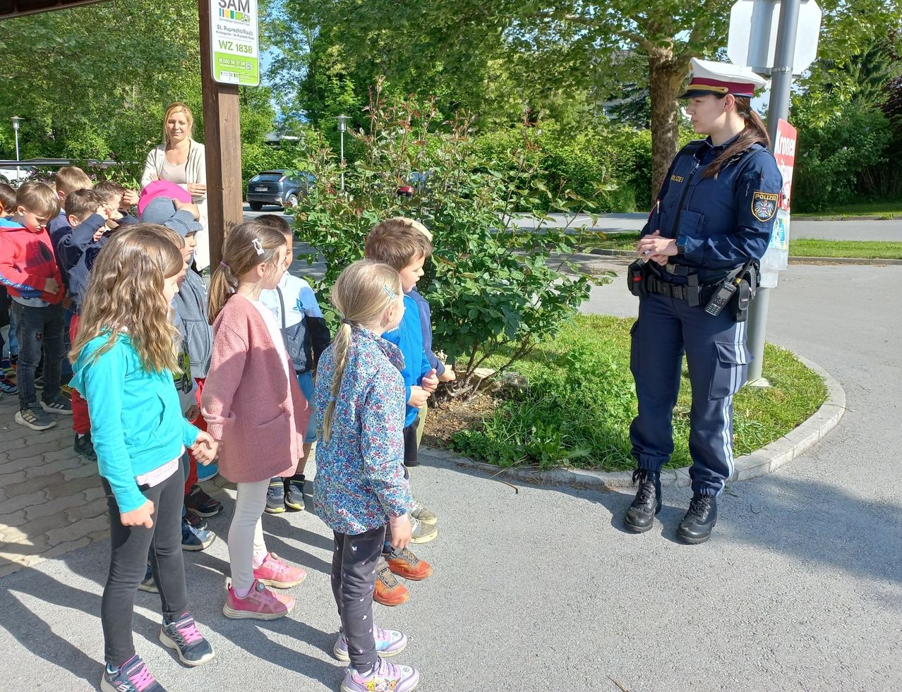 Eine Polizistin erklärt etwas einer Gruppe von Kindern auf dem Bürgersteig. In der Nähe befindet sich ein Schild mit dem Text SAM und einer Frau im Hintergrund.