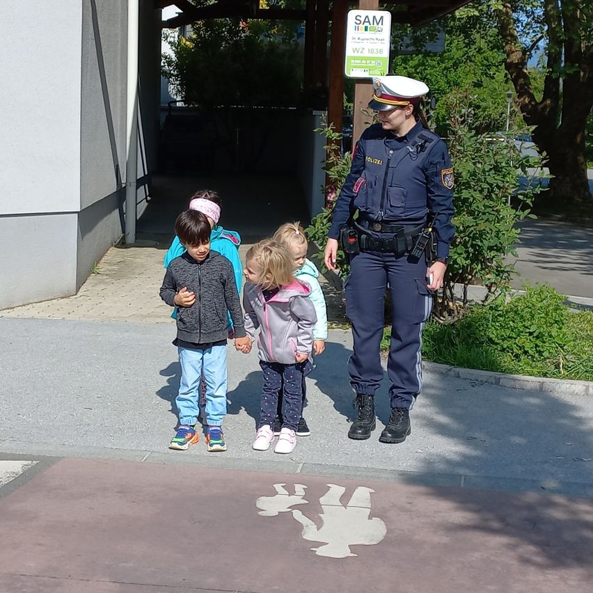 Eine Polizistin steht mit drei Kindern auf einem Bürgersteig in der Nähe eines SAM-Schildes. Die Kinder halten Händchen, und ein Fußgängersymbol ist auf dem Boden gemalt.
