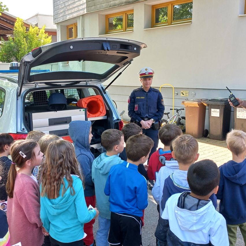 Ein Polizeibeamter steht vor einem Polizeifahrzeug mit offenem Kofferraum, umringt von Kindern, die aufmerksam zuhören. Hinter dem Beamten ist ein Gebäude mit Fenstern und Mülleimern zu sehen.