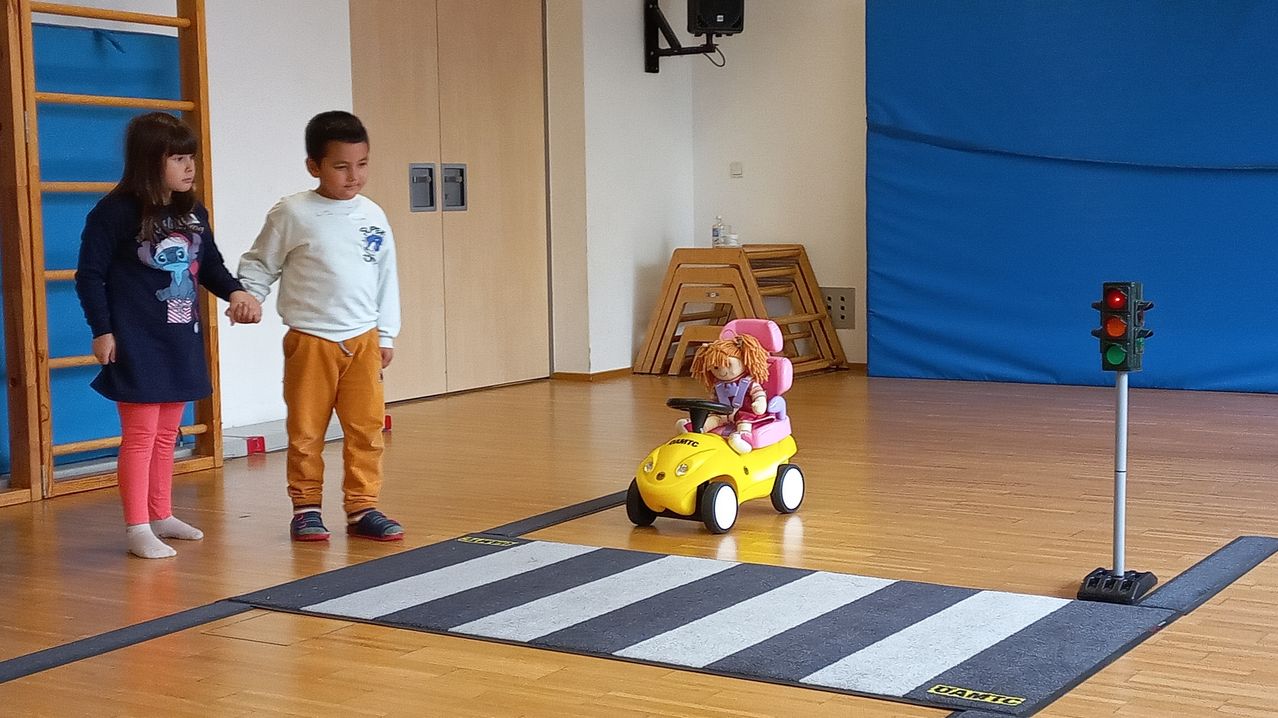Ein kleiner Junge beobachtet ein Mädchen in einem Puppenwagen auf einem Holzboden, mit Schränken und einem Lautsprecher im Hintergrund.
