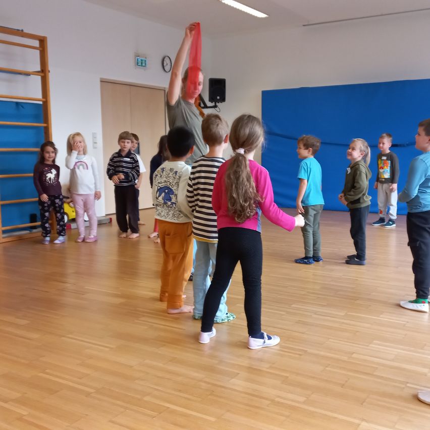 Eine Gruppe von Kindern versammelt sich in einem Raum, in dem eine Frau eine Tanzaktivität mit einem roten Tuch leitet. Die Kinder stehen im Kreis, und der Raum hat Holzböden und weiße Wände.