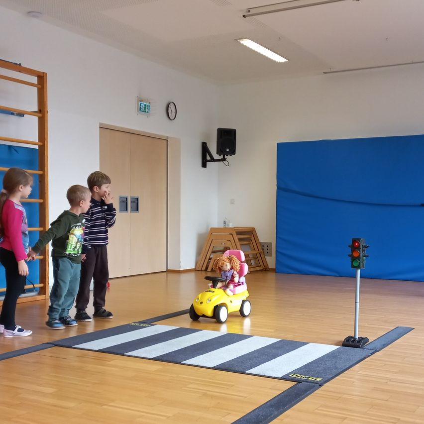 Vier Kinder beobachten, wie ein Spielzeugauto über eine simulierte Straße fährt, mit einer Ampel an der Seite. Der Raum ist mit einem blauen Hintergrund und einer Holzleiter ausgestattet.
