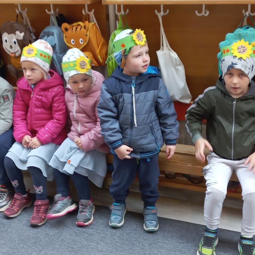 Fünf Kinder sitzen auf einer Bank in einem Raum. Sie tragen Mützen mit Sonnenblumen darauf. Zwei Mädchen tragen rosa Jacken. Ein Junge trägt eine blaue Jacke. Ein anderer Junge trägt eine grüne Jacke.