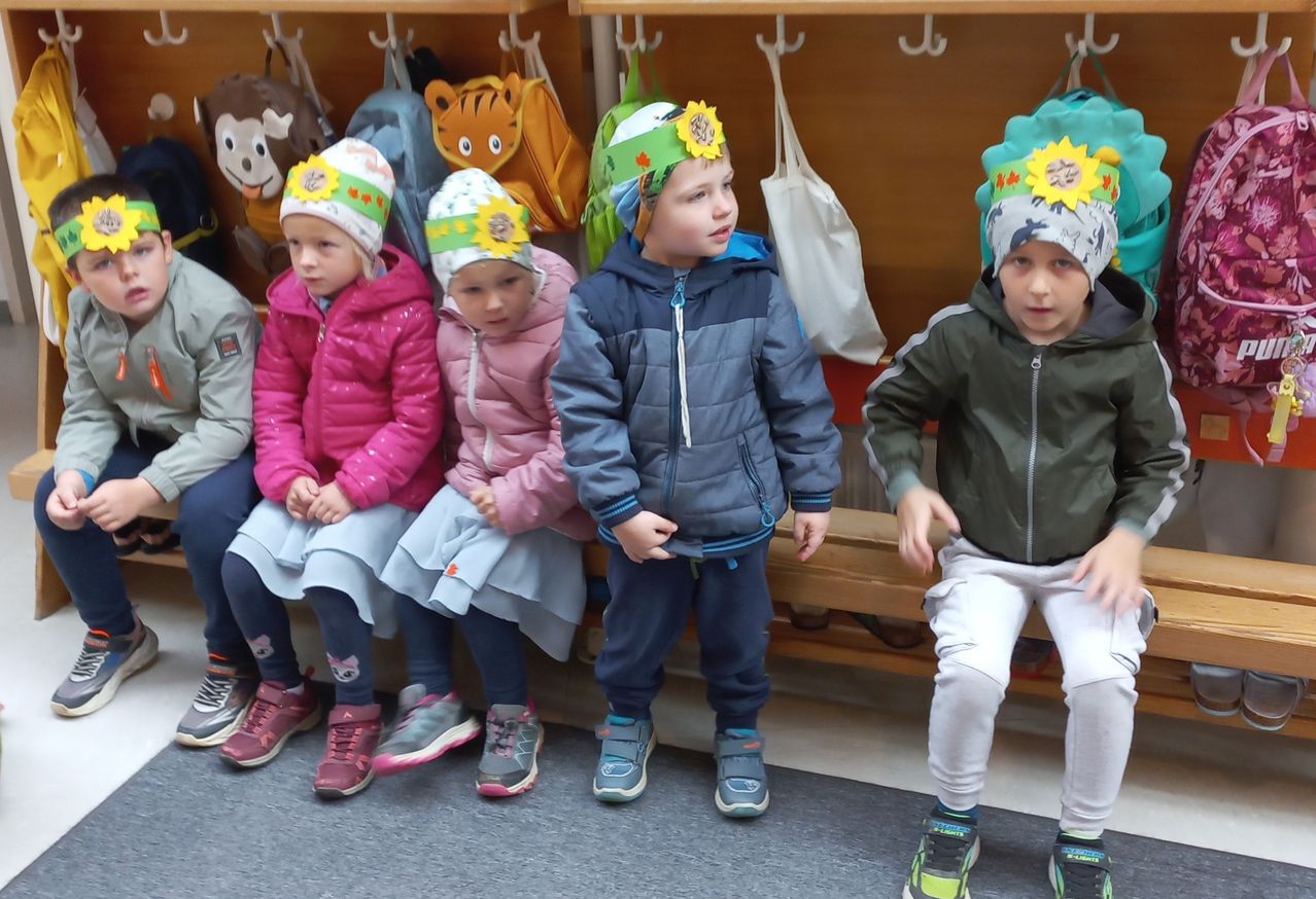 Fünf Kinder sitzen auf einer Bank in einem Raum. Sie tragen Mützen mit Sonnenblumen darauf. Zwei Mädchen tragen rosa Jacken. Ein Junge trägt eine blaue Jacke. Ein anderer Junge trägt eine grüne Jacke.