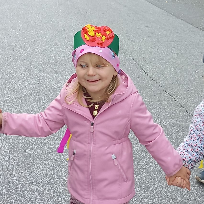 Ein kleines Mädchen in einer rosa Jacke und einem Hut mit einer Blume darauf geht mit einem anderen Kind auf einer Straße.