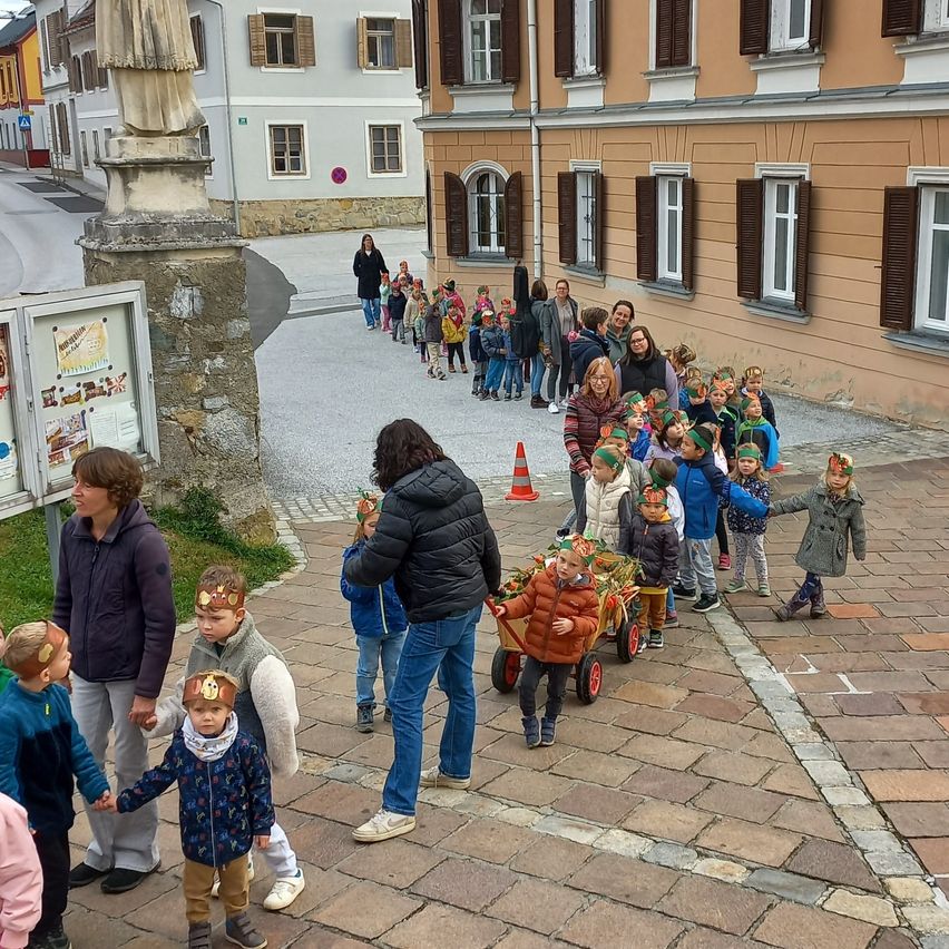 Eine Gruppe von Kindern mit Stirnbändern geht in einer Reihe mit einem Wagen vor einem Gebäude. In der Nähe führen Erwachsene sie.