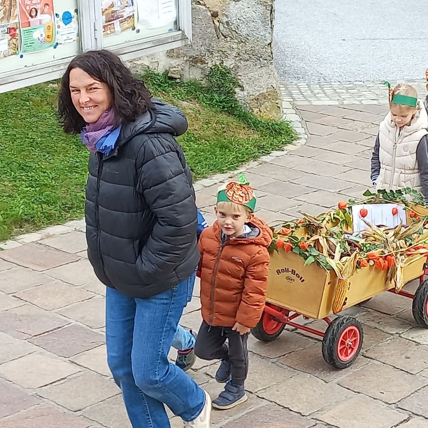 Eine Frau mit lächelndem Gesicht geht mit zwei Kindern auf dem Bürgersteig. Ein Kind hält einen Wagen mit Blumen. Dahinter befindet sich ein Rasenbereich mit einem Gebäude.