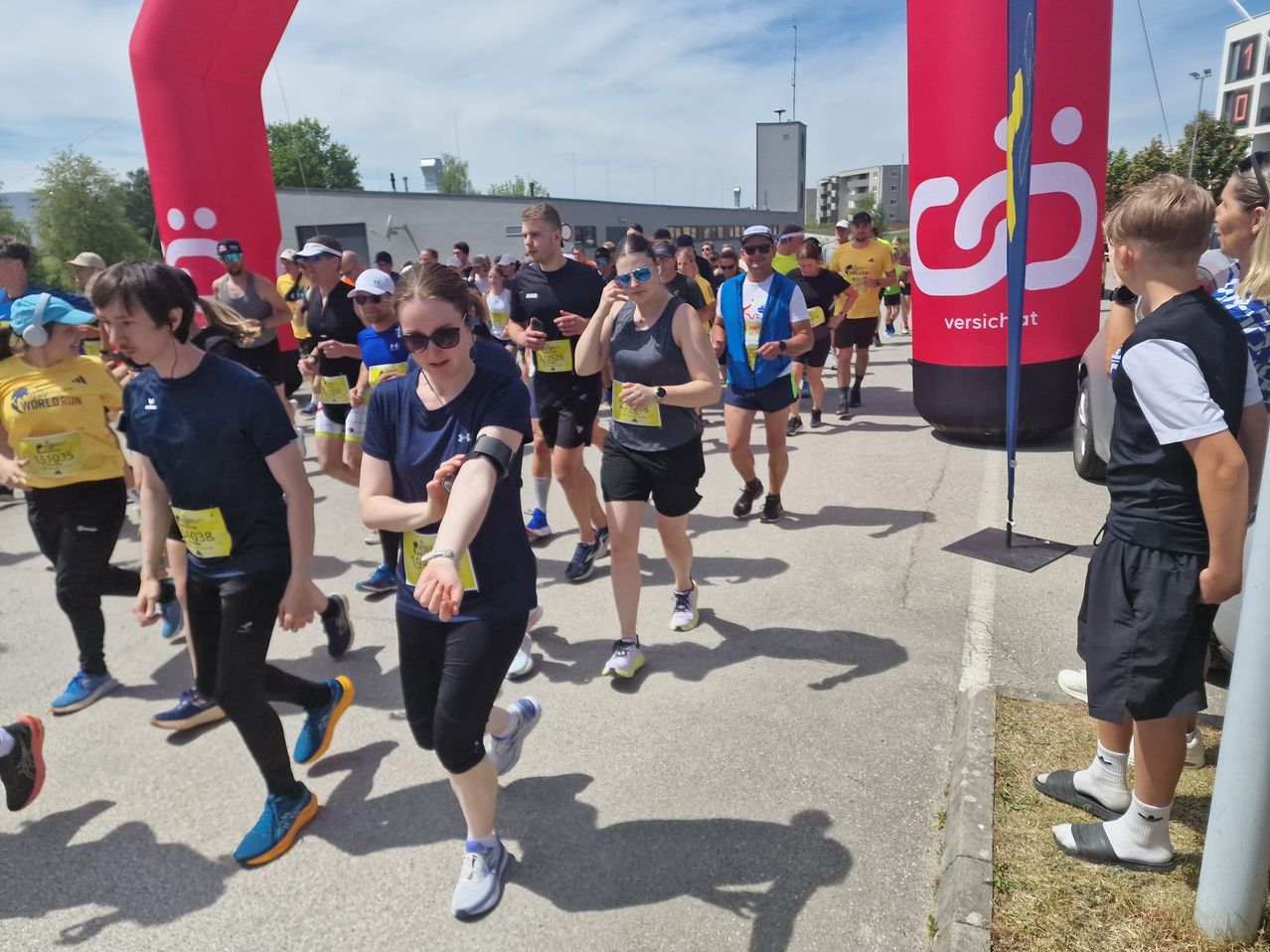 A marathon event with runners in various outfits, including sunglasses, caps, and sneakers. Participants are lined up under a blue sky. The event has red banners on the side.