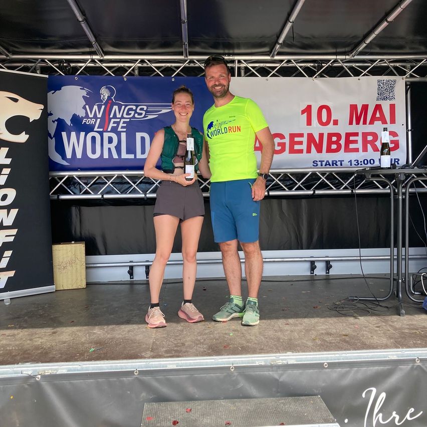 A man and a woman stand on a stage with a bottle of champagne, smiling for a photo. They are at an event for the Wings for Life World Run.