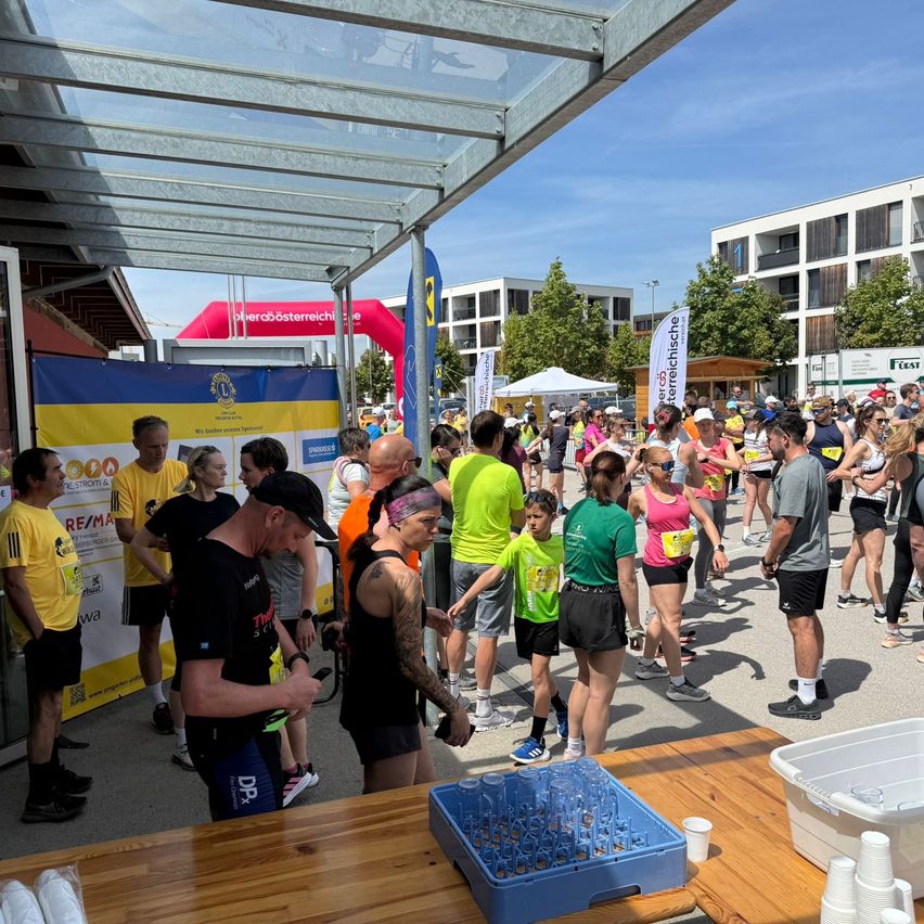Many people wearing various colored jerseys gather outdoors for a marathon. A few are standing, and others are walking. A table with cups and plates is nearby.