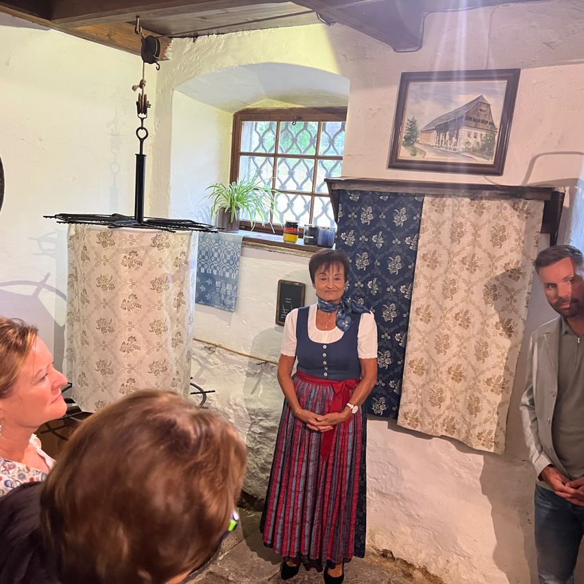 Eine Frau in traditioneller bayerischer Kleidung steht in einem Raum mit Fenster, Gemälde und hängender Lampe.