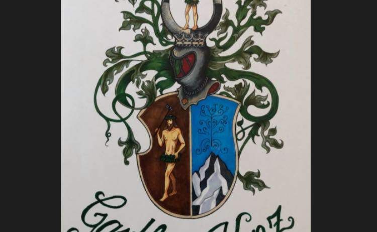 Ein illustriertes Wappen mit dem Namen Gasthaus Kantz unten. Es zeigt einen Mann, der einen Hammer über einem Schild hält, mit einem Berg auf dem Schild. Grüne Blätter und Reben umgeben den Schild.