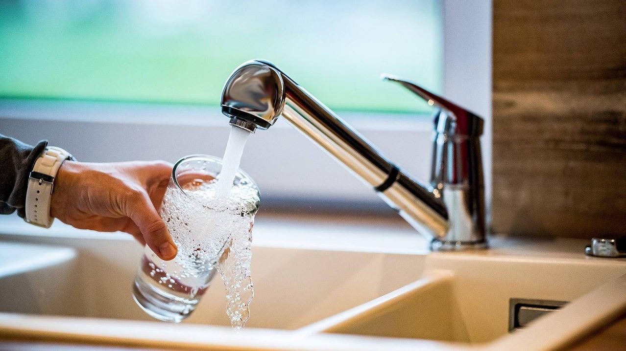 Eine Hand füllt ein Glas mit Wasser aus einem silbernen Wasserhahn. Das Becken ist aus beigem Material.