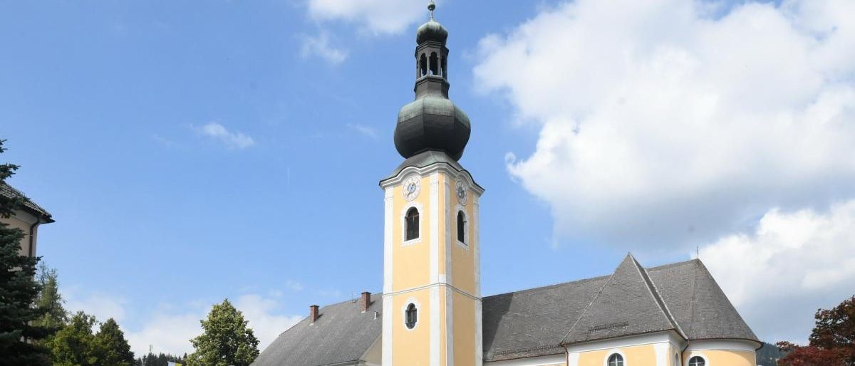 Eine gelbe Kirche mit einem grauen Dach und einem hohen Turm mit einer Uhr. Die Kirche hat viele Fenster und ist von Büschen und Bäumen umgeben. Eine weiße Fahne befindet sich links.