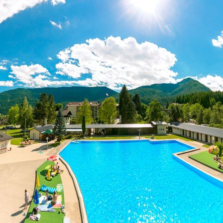 Eine Vogelperspektive auf einen großen Swimmingpool, umgeben von üppigem Grün und Bergen unter einem klaren blauen Himmel.