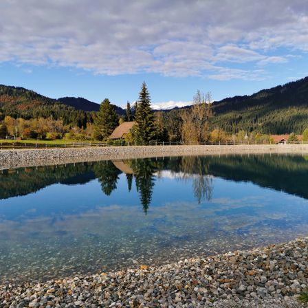 Ein ruhiger See, der die Berge und Bäume unter einem teilweise bewölkten Himmel widerspiegelt. Steiniger Uferrand mit Kies und Bäumen im Hintergrund.