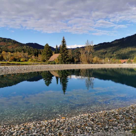 Ein ruhiger See, der die Berge und Bäume unter einem teilweise bewölkten Himmel widerspiegelt. Steiniger Uferrand mit Kies und Bäumen im Hintergrund.