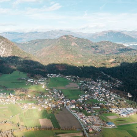 Eine Luftaufnahme eines Dorfes, umgeben von üppigen grünen Feldern und bewaldeten Bergen unter einem teilweise bewölkten Himmel.