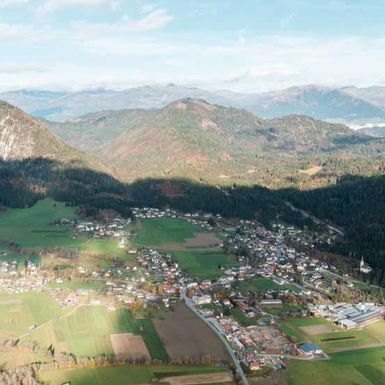 Eine Luftaufnahme eines Dorfes, umgeben von üppigen grünen Feldern und bewaldeten Bergen unter einem teilweise bewölkten Himmel.