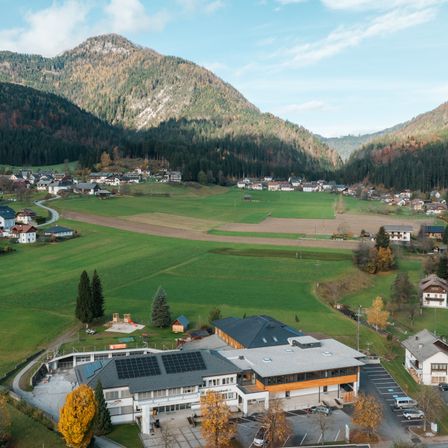Eine Luftaufnahme eines Dorfes mit einem Schulgebäude, umgeben von grünen Feldern, Bäumen und Bergen unter einem blauen Himmel mit einigen Wolken.