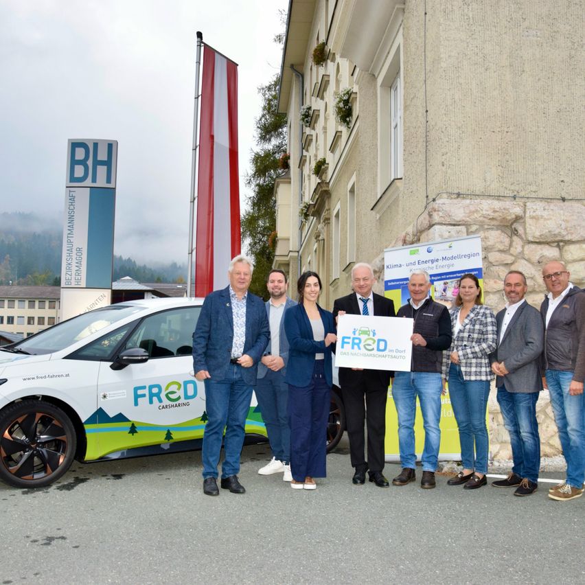 Eine Gruppe von Personen steht vor einem Gebäude mit einem grünen Auto, das mit 'FRED Carsharing' beschriftet ist. Ein Banner mit einer Flagge und einem Logo ist sichtbar.