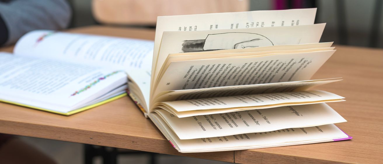 Ein Stapel offener Bücher und Notizbücher liegt auf einem Schreibtisch in einem Klassenzimmer. Die Bücher sind ausgebreitet, wobei eines von ihnen geöffnet ist und eine Zeichnung zeigt.