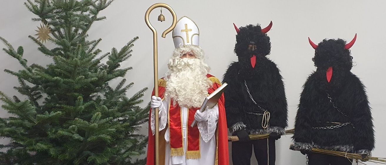 Ein Mann in Santa-Claus-Kostüm hält einen Stab, neben ihm stehen zwei Teufel in schwarzen Kostümen, alle vor einem Weihnachtsbaum.