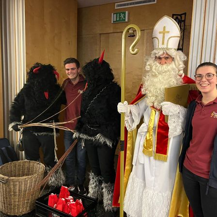 Eine Gruppe von Menschen, die als Teufel verkleidet sind, und ein Mann, der als St. Nikolaus verkleidet ist, stehen zusammen mit einer lächelnden Frau vor der Kamera.