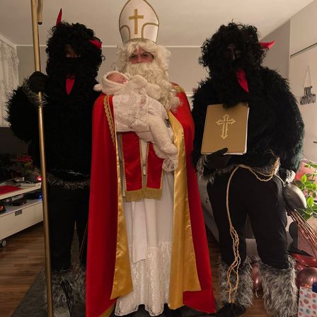 Ein Mann, der als Weihnachtsmann verkleidet ist, hält ein Baby, während zwei Personen in Teufelskostümen neben ihm stehen, eine hält ein Buch mit einem Kreuz darauf. Die Umgebung ist drinnen, mit Möbeln und Dekorationen im Hintergrund.