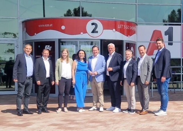 Sieben Personen stehen vor einem Gebäudeeingang, möglicherweise für ein Foto. Sie tragen formelle Kleidung. Ein großes Schild mit der Nummer '2' befindet sich an der Wand.