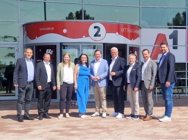 Sieben Personen stehen vor einem Gebäudeeingang, möglicherweise für ein Foto. Sie tragen formelle Kleidung. Ein großes Schild mit der Nummer '2' befindet sich an der Wand.