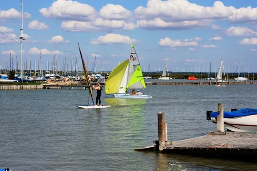 Eine Person segelt mit einem Windsurf-Board in der Nähe eines Stegs, mit einem weiteren Windsurf-Boot im Hintergrund und einem bewölkten Himmel.