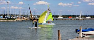 A person is sailing a windsurf board near a dock, with another windsurf boat in the background, and a cloudy sky.