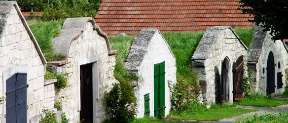 Ein Dorf mit Steingebäuden und roten Dächern, umgeben von Bäumen, mit einem weißen Kirchturm in der Ferne.
