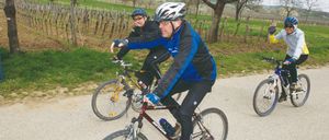 Two cyclists ride their bikes on a gravel road, with a man wearing a helmet and holding a bottle in the front. Another cyclist follows behind. They are surrounded by a vineyard with bare trees and a grassy field.
