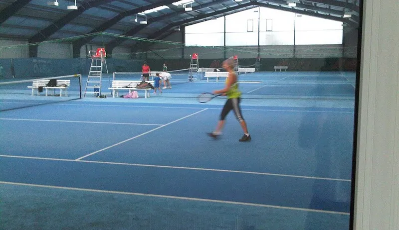 Eine überdachte Tennishalle mit einer Frau, die Tennis spielt. Bänke, Netze und Menschen sind zu sehen. Zwei Schiedsrichter stehen auf Leitern in der Nähe des Platzes. Der Platz hat einen blauen Boden und weiße Markierungen.