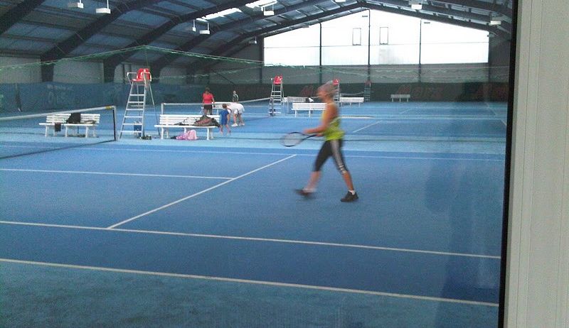 Eine überdachte Tennishalle mit einer Frau, die Tennis spielt. Bänke, Netze und Menschen sind zu sehen. Zwei Schiedsrichter stehen auf Leitern in der Nähe des Platzes. Der Platz hat einen blauen Boden und weiße Markierungen.