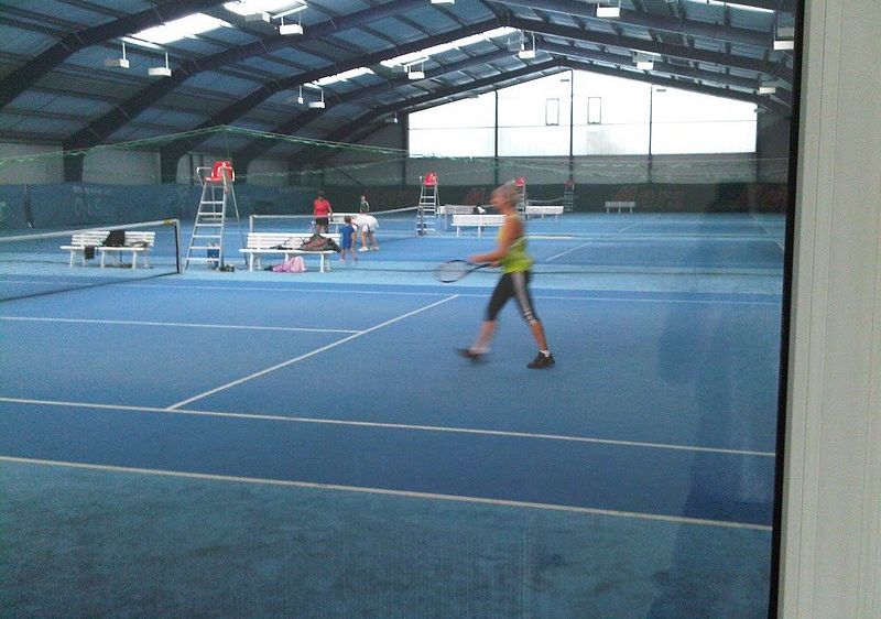Eine überdachte Tennishalle mit einer Frau, die Tennis spielt. Bänke, Netze und Menschen sind zu sehen. Zwei Schiedsrichter stehen auf Leitern in der Nähe des Platzes. Der Platz hat einen blauen Boden und weiße Markierungen.
