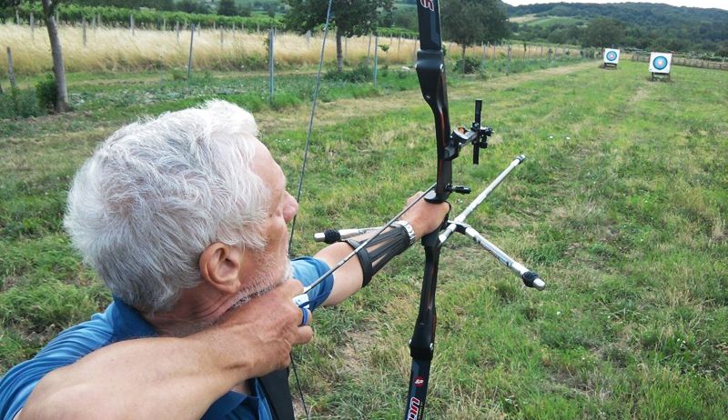 Ein älterer Mann zielt mit einem Bogen in einem offenen Feld auf zwei Ziele in der Ferne.