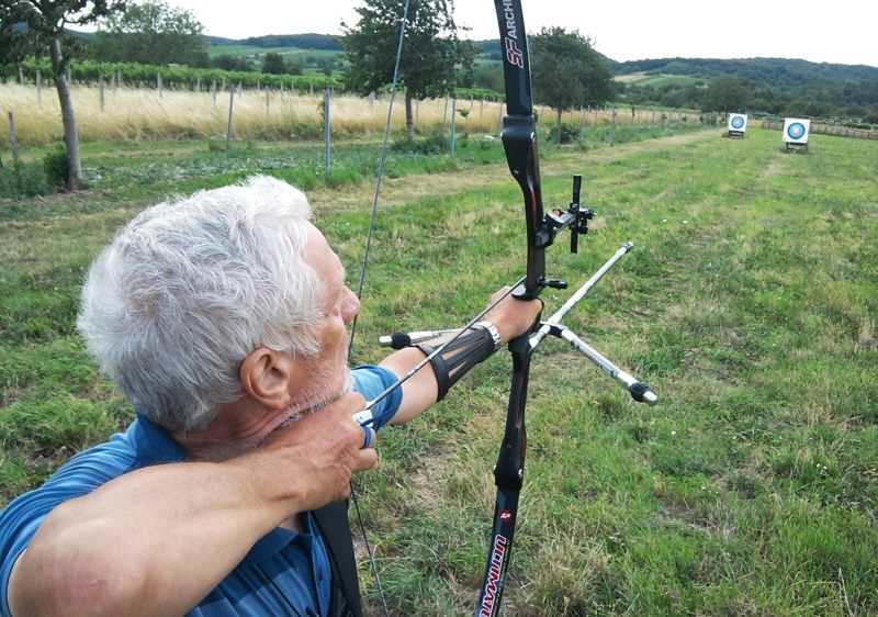 Ein älterer Mann zielt mit einem Bogen in einem offenen Feld auf zwei Ziele in der Ferne.
