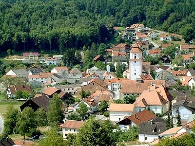 Luftaufnahme eines Dorfes mit zahlreichen Häusern, überwiegend mit roten Dächern, umgeben von üppigem Grün und Bäumen. Im Zentrum sticht ein weißer Turm hervor.