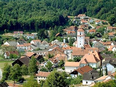 Luftaufnahme eines Dorfes mit zahlreichen Häusern, überwiegend mit roten Dächern, umgeben von üppigem Grün und Bäumen. Im Zentrum sticht ein weißer Turm hervor.