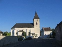 Eine weiße Kirche mit einem grauen Dach und einem hohen Turm steht am Straßenrand. Bäume und ein geparktes Auto sind in der Nähe.